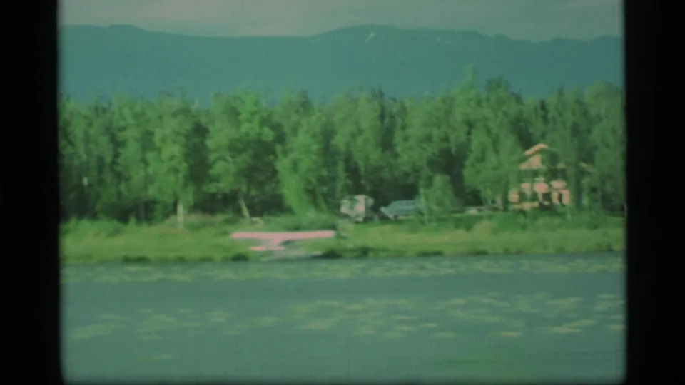1977 Alaska lakeshore frame with evergreens, a small pink seaplane on the water, vehicles, and a house near the treeline.