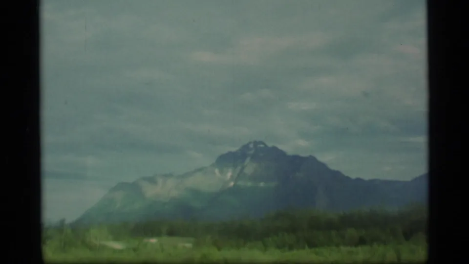 1977 Alaska 8mm frame showing a snow-marked mountain beyond forest and low foreground blur beneath heavy clouds.