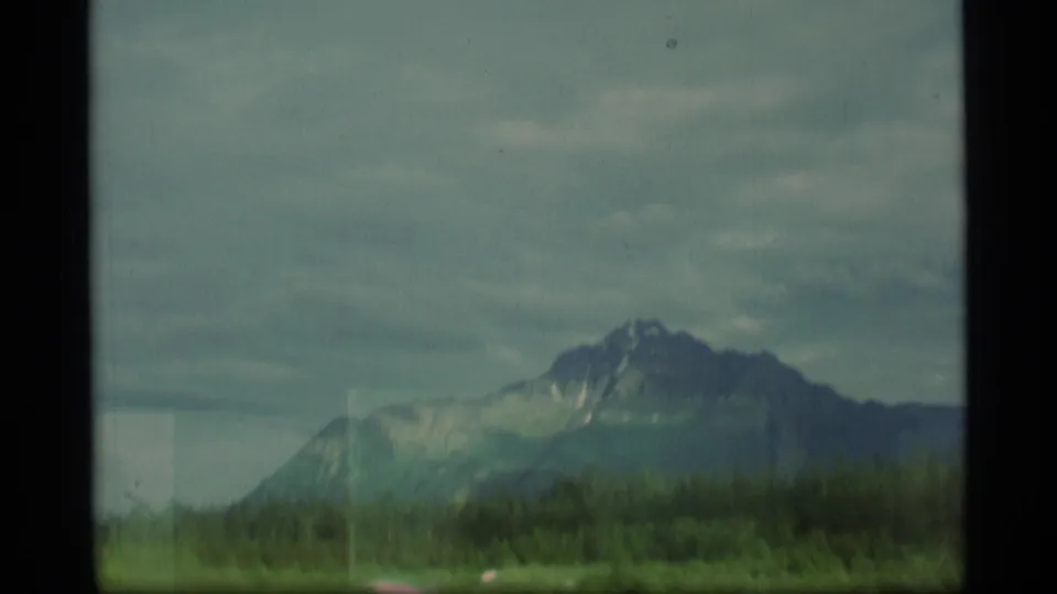 1977 Alaska 8mm frame with a snow-marked mountain, evergreen horizon, cloudy sky, and soft home-movie exposure.