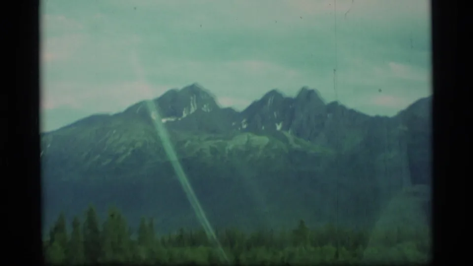 1977 Alaska home-movie frame with a mountain range, forested lower slopes, window framing, and soft green-blue 8mm color.