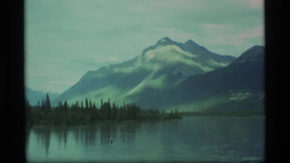 1977 Alaska 8mm frame showing a still lake, reflected treeline, and a mountain slope under soft, hazy light.