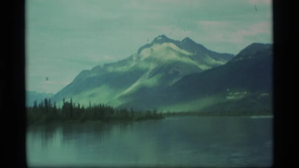 1977 Alaska travel frame showing a broad lake foreground, dark evergreen shore, and layered mountain slopes in soft 8mm color.