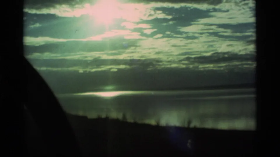 1977 Alaska 8mm frame with sunlit clouds, reflected light on water, and a dark lakeshore silhouette.