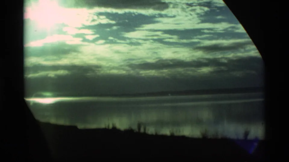 1977 Alaska 8mm frame showing a bright sunlit sky, layered clouds, reflective water, and dark foreground land.