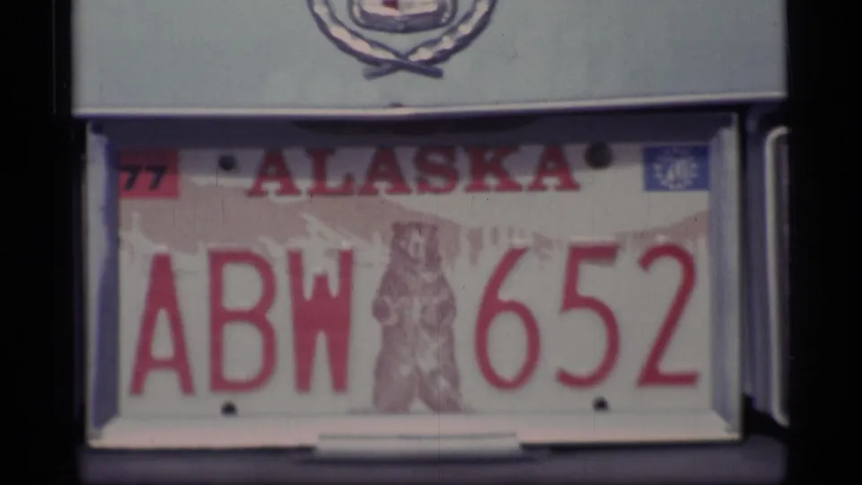 1977 Alaska license plate ABW 6552 with bear emblem, mounted beneath a Cadillac crest in archival 8mm footage.