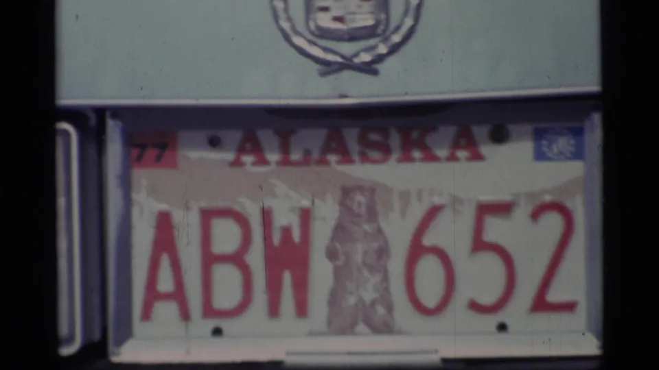 Close 8mm frame of the 1977 Alaska ABW 6552 plate with the bear graphic and Cadillac emblem visible above it.