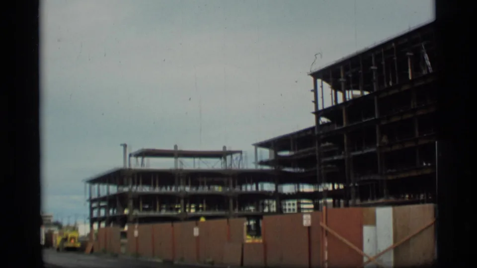 1977 Alaska home-movie frame showing multiple steel-frame sections of a federal building site behind temporary fencing.