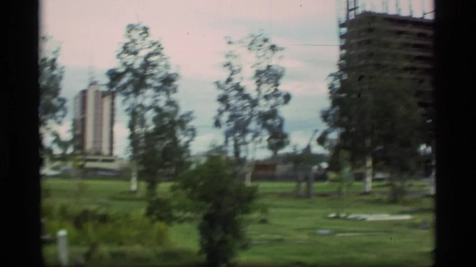 Soft 8mm view of an Alaska urban green space with trees, distant buildings, and part of the construction site at the right edge.