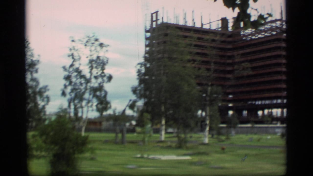 1977 Alaska 8mm frame showing a large high-rise structure under construction beyond a grassy area with trees.