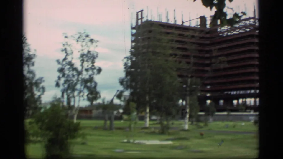 1977 Alaska 8mm frame showing a large high-rise structure under construction beyond a grassy area with trees.