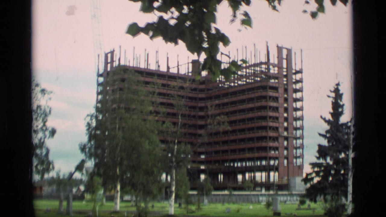 Unfinished high-rise building seen through trees with construction equipment nearby.