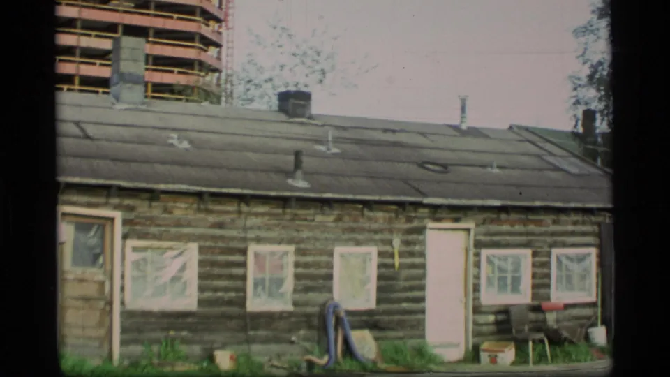 1977 Alaska frame showing a weathered log cabin in the foreground with the high-rise construction visible behind it.