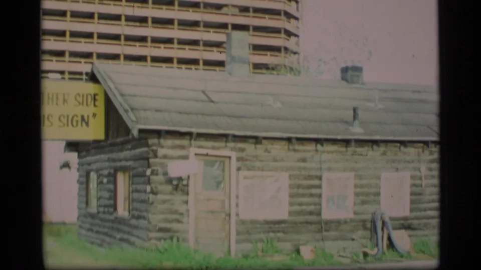 1977 Alaska 8mm frame showing a log cabin corner, a partial roadside sign, and the unfinished high-rise in the background.