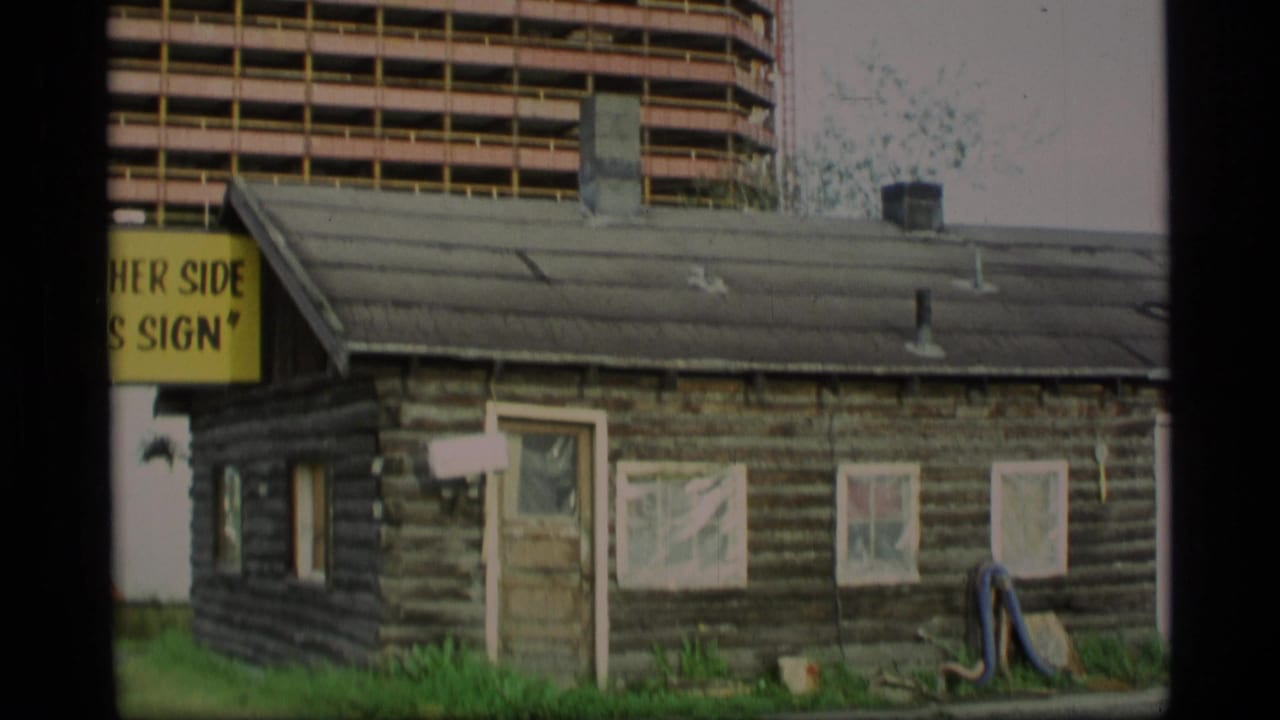 Weathered log cabin with a partial yellow sign and a modern high-rise behind it.