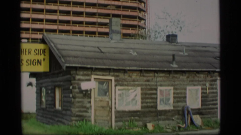 1977 Alaska 8mm frame showing a small log cabin in front of a taller modern building, with a partial yellow sign at the left edge.