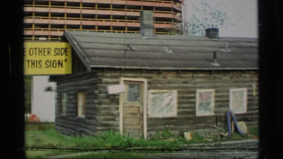 1977 Alaska home-movie frame of a dark log cabin, yellow sign reading part of “THE OTHER SIDE OF THIS SIGN,” and a modern building behind.