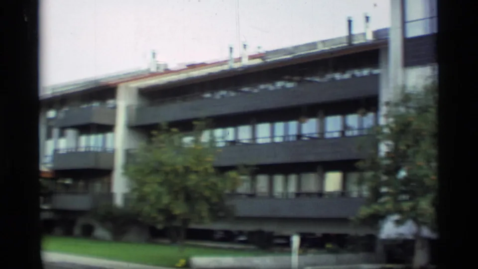 1977 Alaska 8mm frame showing a multi-story modern apartment building with dark balconies, pale columns, lawn, and trees.