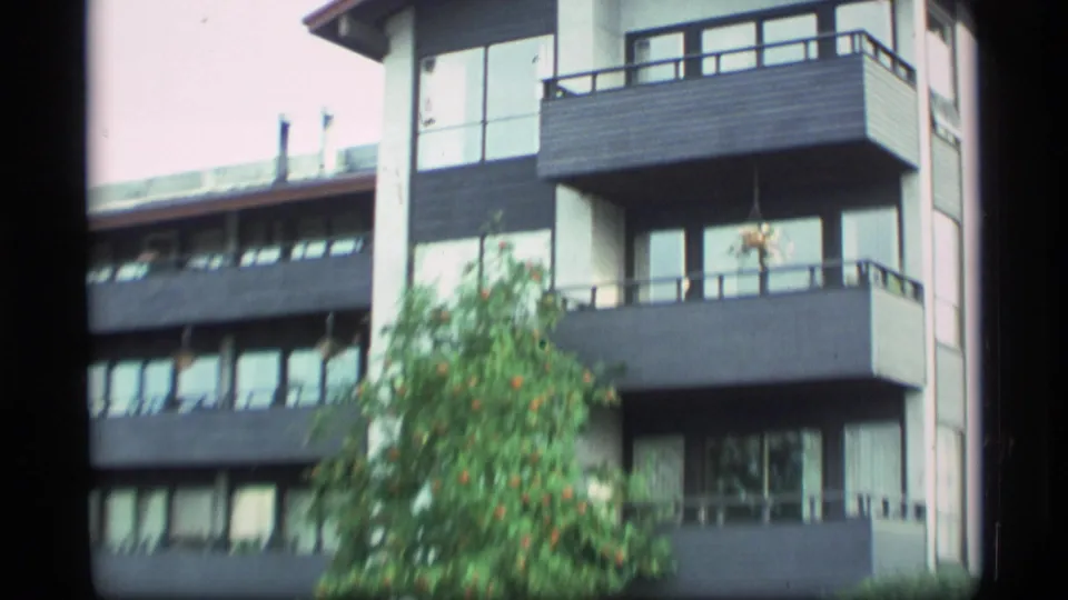 1977 Alaska home-movie frame showing the corner of a modern residential building with stacked balconies, windows, and a tree in the foreground.