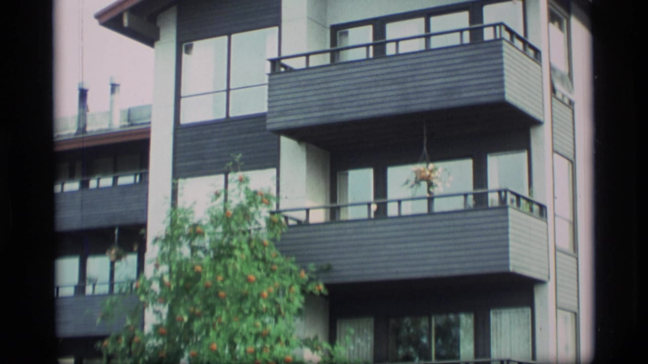 1977 Alaska home-movie frame showing a modern apartment exterior with stacked balconies, windows, and a tree in soft 8mm color.