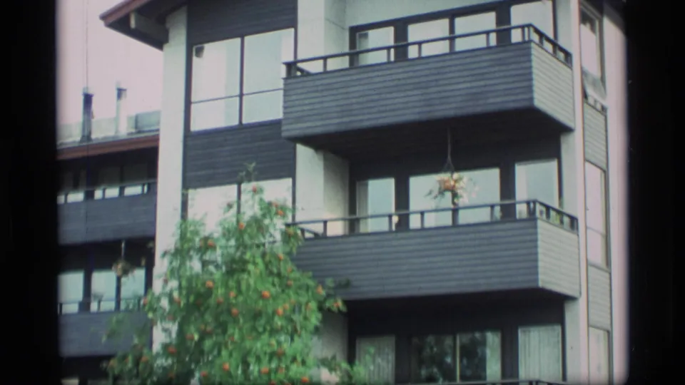 1977 Alaska home-movie frame showing a modern apartment exterior with stacked balconies, windows, and a tree in soft 8mm color.