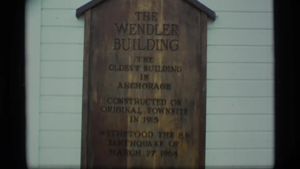 1977 Alaska frame of the wooden Wendler Building sign on a pale exterior wall, with dark film borders and soft focus.