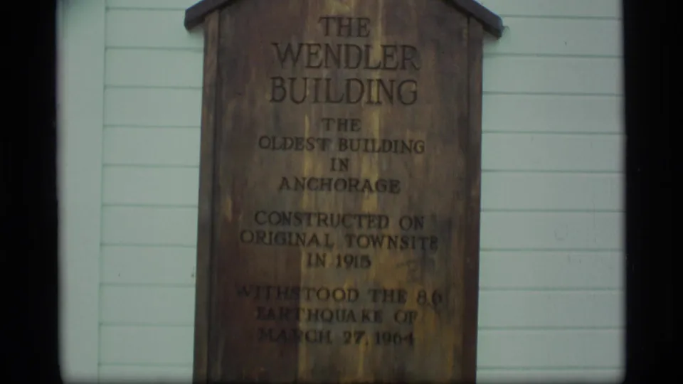 Close 8mm frame of the weathered wooden Wendler Building sign, with readable lines about Anchorage and the 1964 earthquake.