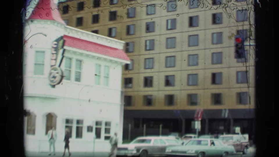 1970s Alaska street view showing the white corner club building, vintage cars, pedestrians, traffic signal, and a larger downtown building behind.
