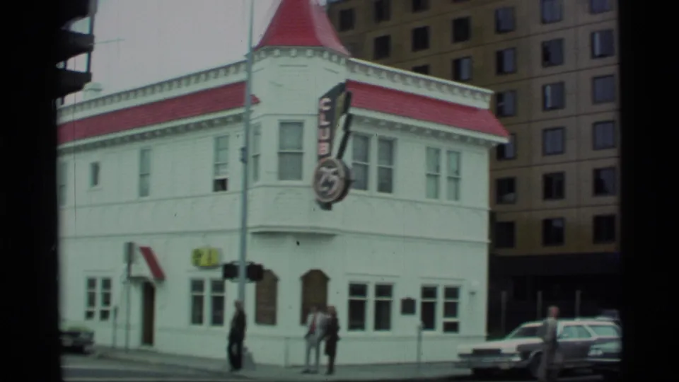 Wide 1977 Alaska 8mm frame of the corner building with CLUB 75 sign, red roof, pedestrians at the sidewalk, and parked or passing cars.