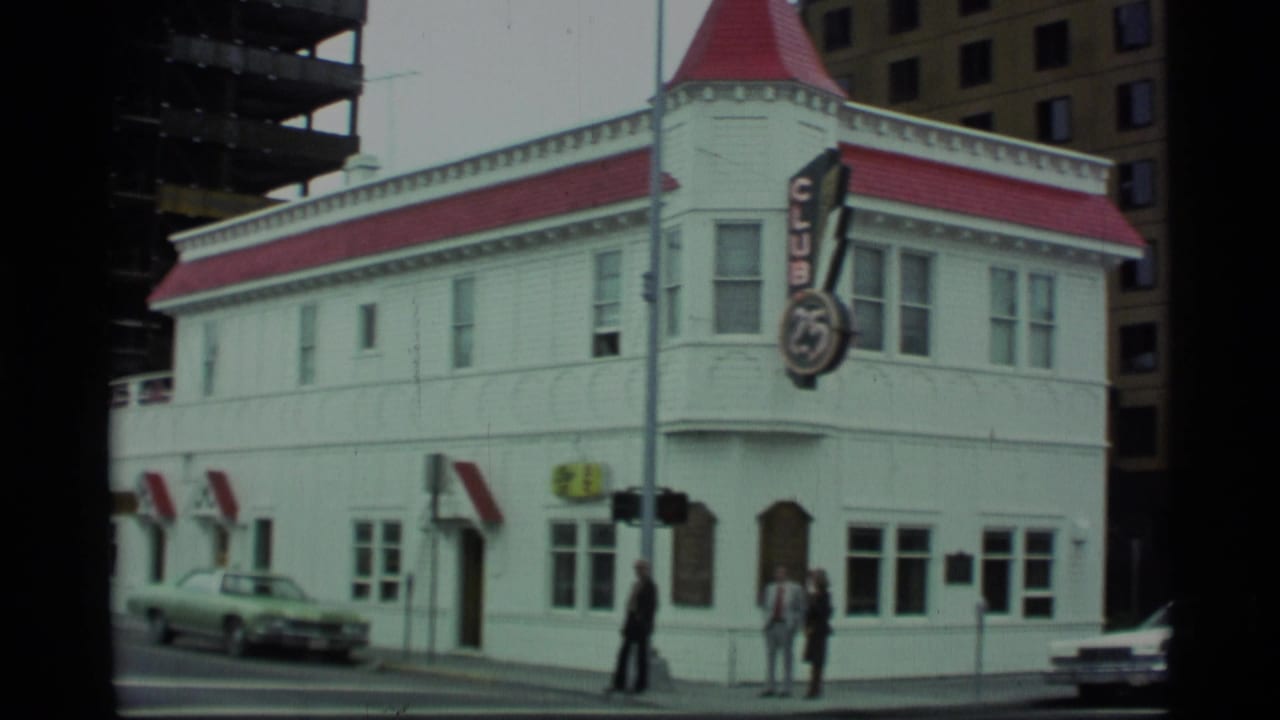 1977 Alaska street view of a white Club 25 building with red roof trim, pedestrians on the sidewalk, parked cars, and construction nearby.
