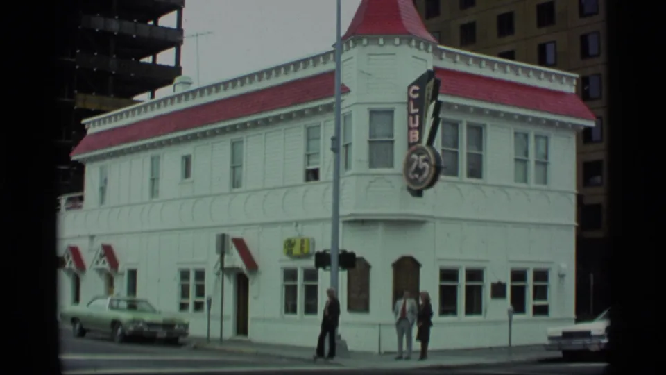 1977 Alaska view of the Club 25 corner facade, with pedestrians near the entrance, a parked car, and taller buildings behind.