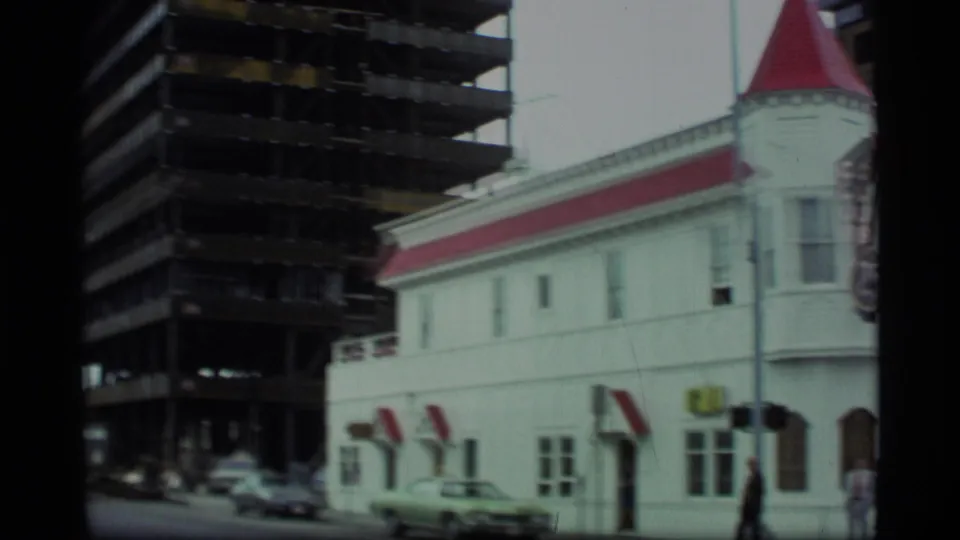 1977 Alaska street-level pan showing the Club 25 building partly beside a dark high-rise frame under construction.