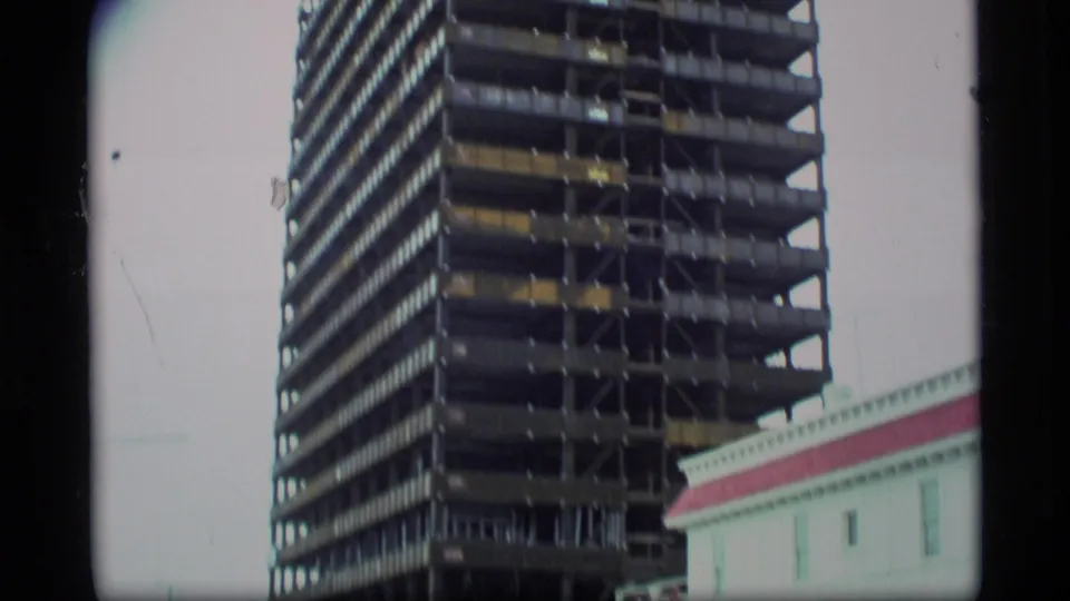1977 Alaska view of a high-rise structure under construction, with the red-trimmed white Club 25 building partly visible at frame edge.