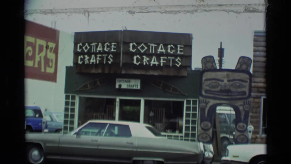 1977 Alaska street view with Cottage Crafts signage, a totem-style arch feature, storefront windows, and 1970s cars parked along the curb.