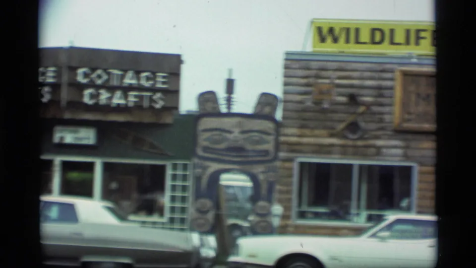 1977 Alaska frame showing the Cottage Crafts sign, part of the Wildlife Museum facade, a totem-style feature, and cars parked outside.