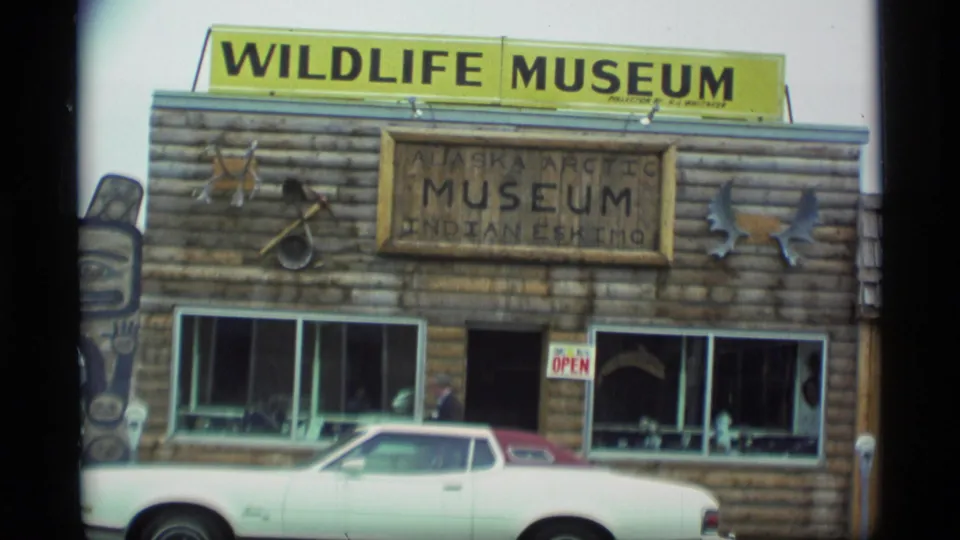 1977 Alaska home-movie frame showing the Wildlife Museum facade, Alaska Arctic Museum sign, antlers, snowshoes, display windows, and a parked car.