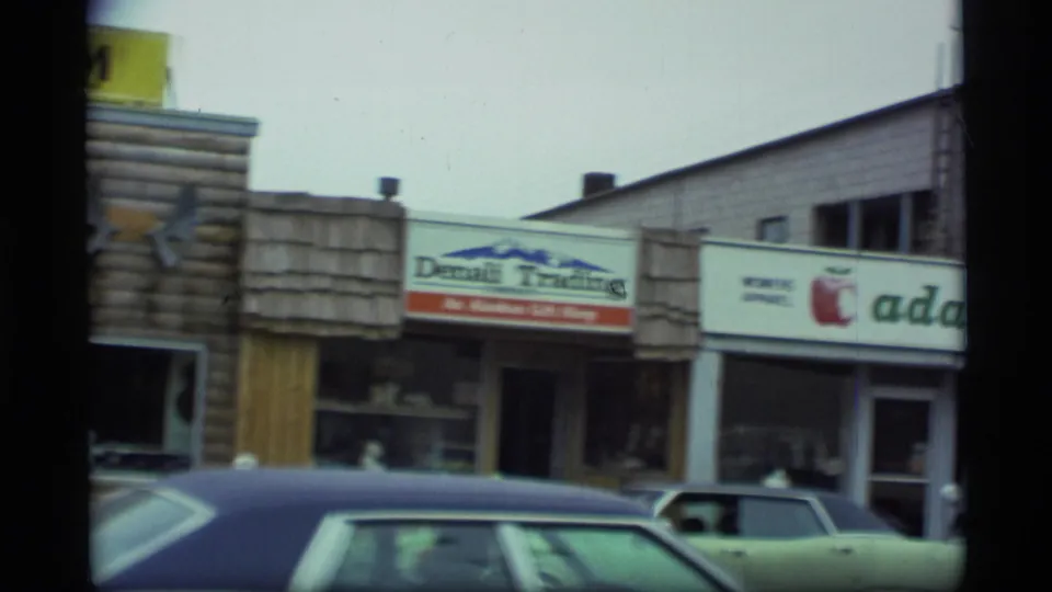 1977 Alaska storefront row with part of the museum facade, Denali Trading sign, women's apparel sign, and parked cars in front.