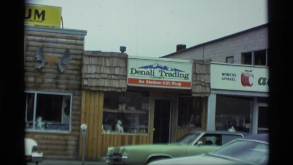 1977 Alaska street scene centered on Denali Trading, a neighboring women's apparel storefront, museum facade edge, and 1970s parked vehicles.
