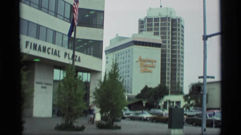 1977 Anchorage urban view showing Financial Plaza lettering, flags, parked cars, and the Anchorage Western Hilton high-rise in the background.
