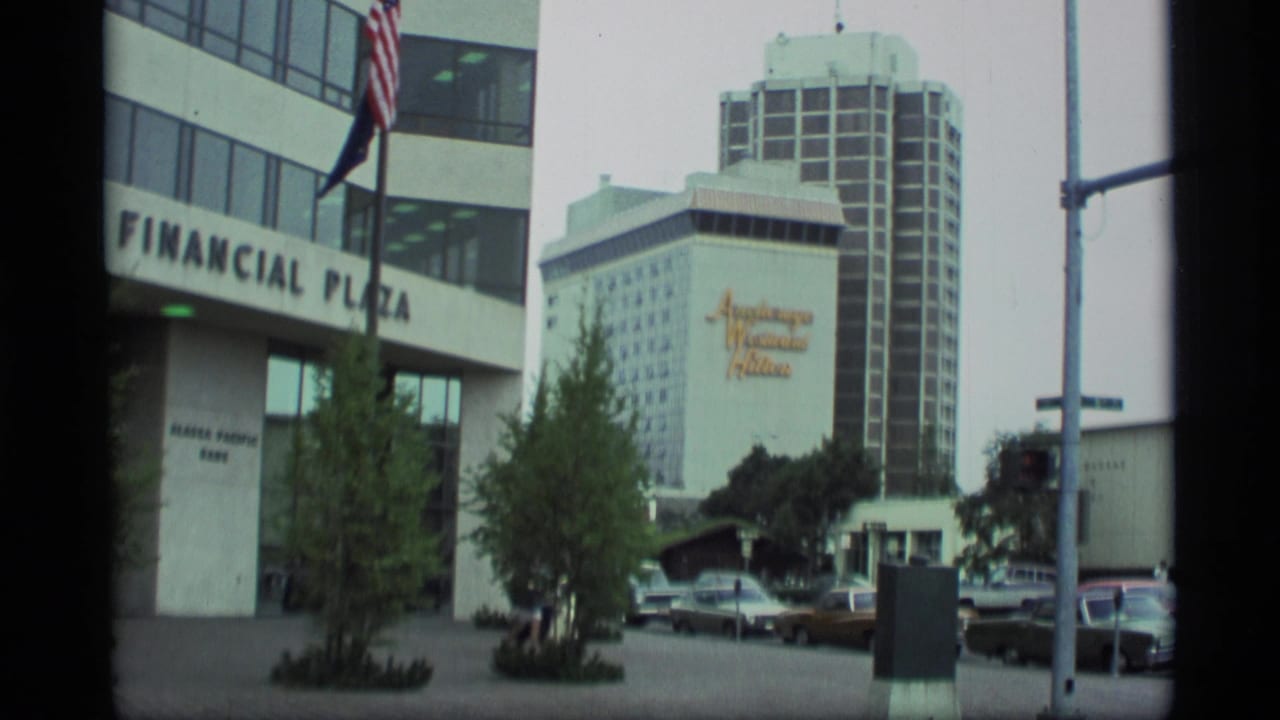 Downtown Anchorage street view with Financial Plaza, hotel signage, parked cars, trees, and an American flag.