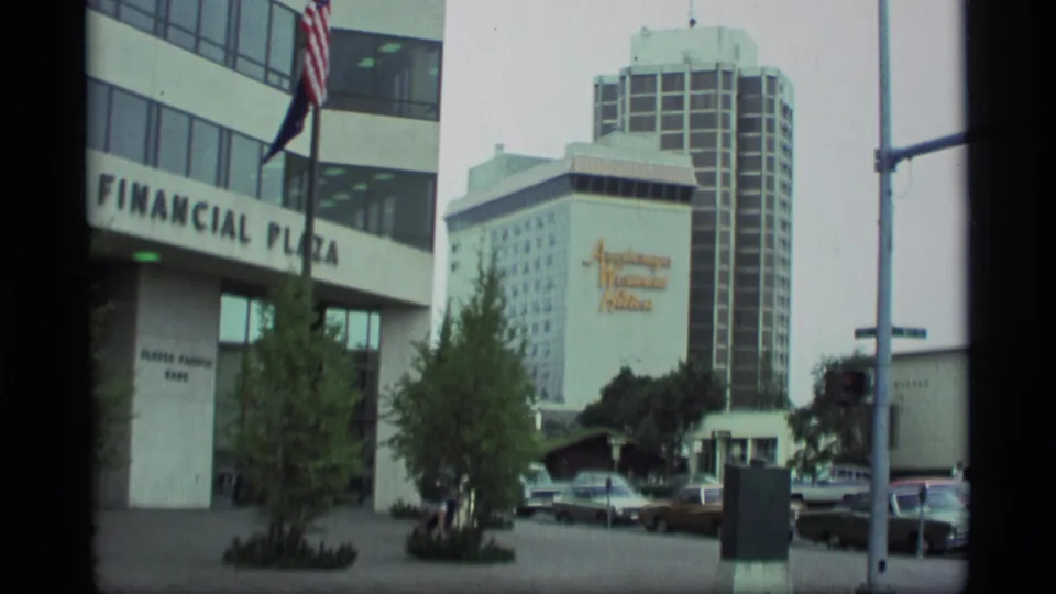 1977 Anchorage street scene showing Financial Plaza at left, Anchorage Westin Hilton signage in the background, parked cars, trees, and an American flag.
