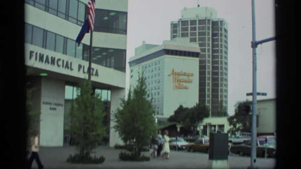 1977 downtown Anchorage view with Financial Plaza, the Anchorage Westin Hilton sign, parked 1970s cars, trees, and pedestrians near the sidewalk.