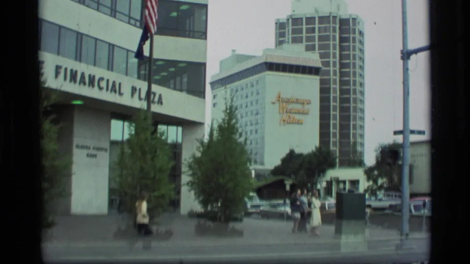 1977 Anchorage street footage showing Financial Plaza, hotel signage, a row of parked cars, trees, and pedestrians moving through the frame.