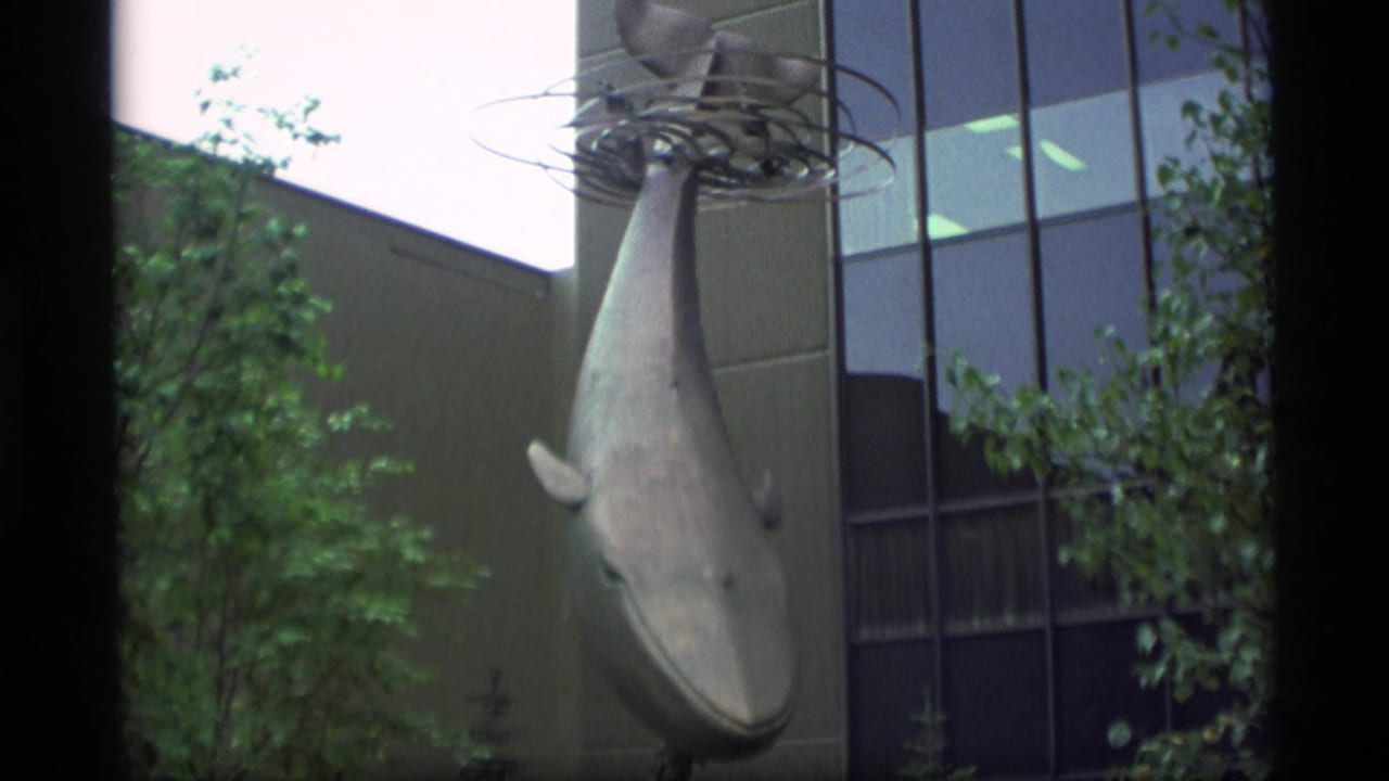 Closer view of the whale sculpture beside glass windows and greenery.