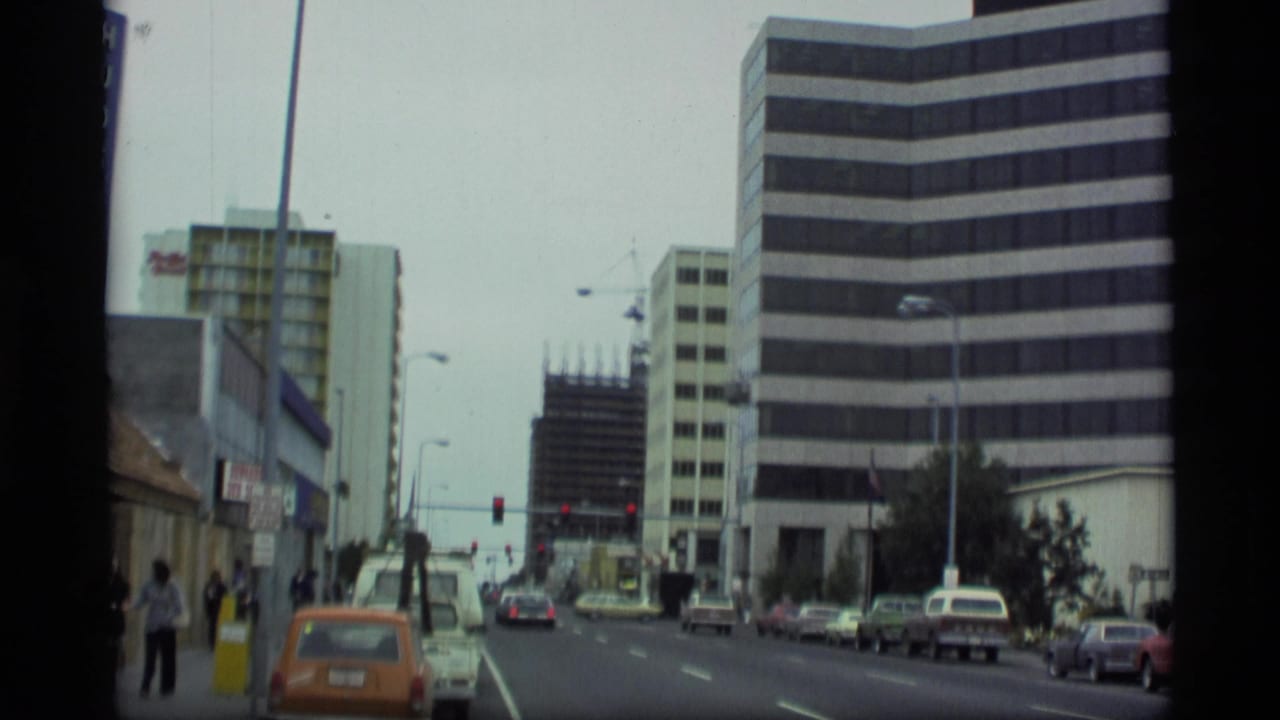 Urban street with older cars, pedestrians, traffic lights, and a striped office building.
