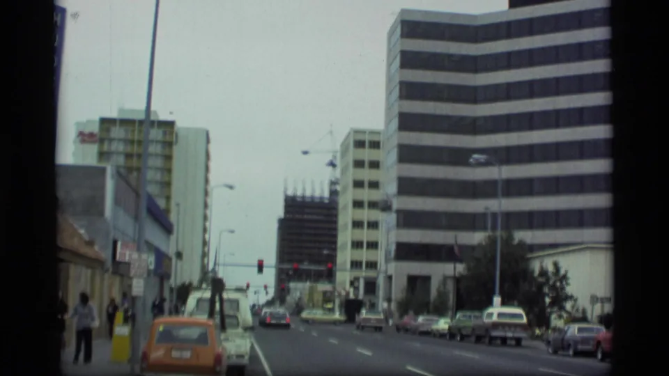 1977 Alaska street scene with older cars, pedestrians on the sidewalk, traffic signals, a striped office building, and mid-rise commercial buildings.