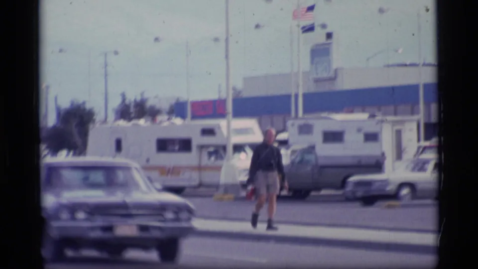 1977 Alaska street-side view with a man walking across the foreground, parked RVs, older cars, poles, and a commercial building with visible signage.