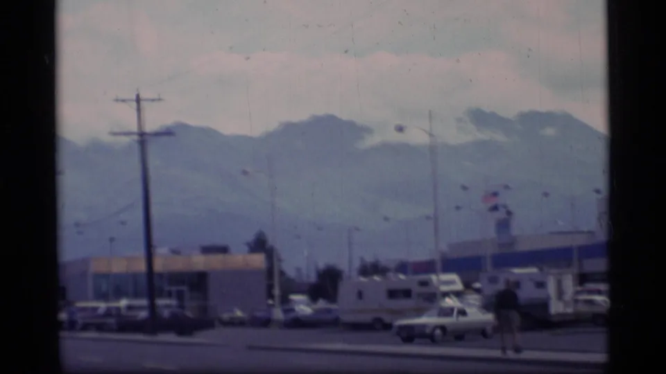 1977 Alaska city-edge view with distant mountains, parked vehicles, RVs, utility poles, low commercial buildings, and a pedestrian near the roadside.