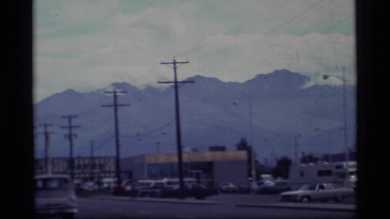 1977 Alaska 8mm street view with utility poles, parked cars, commercial buildings, and distant mountains.