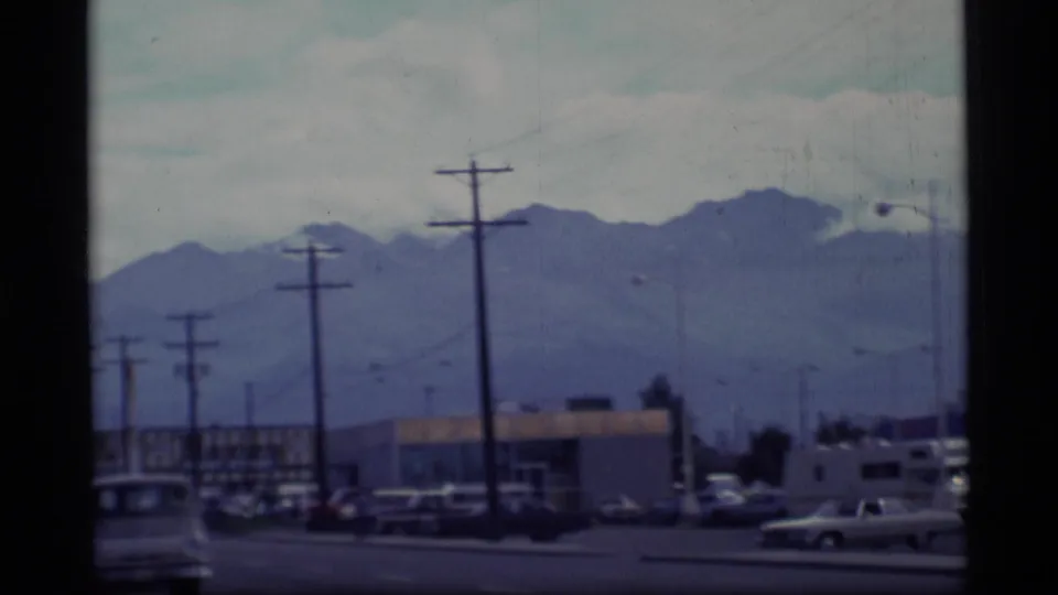 1977 Alaska 8mm street view with utility poles, parked cars, commercial buildings, and distant mountains.