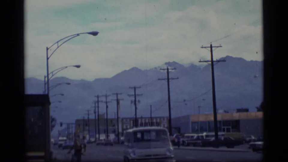 Drive-by 1977 Alaska street view with street lamps, utility poles, cars, storefronts, and mountains behind them.